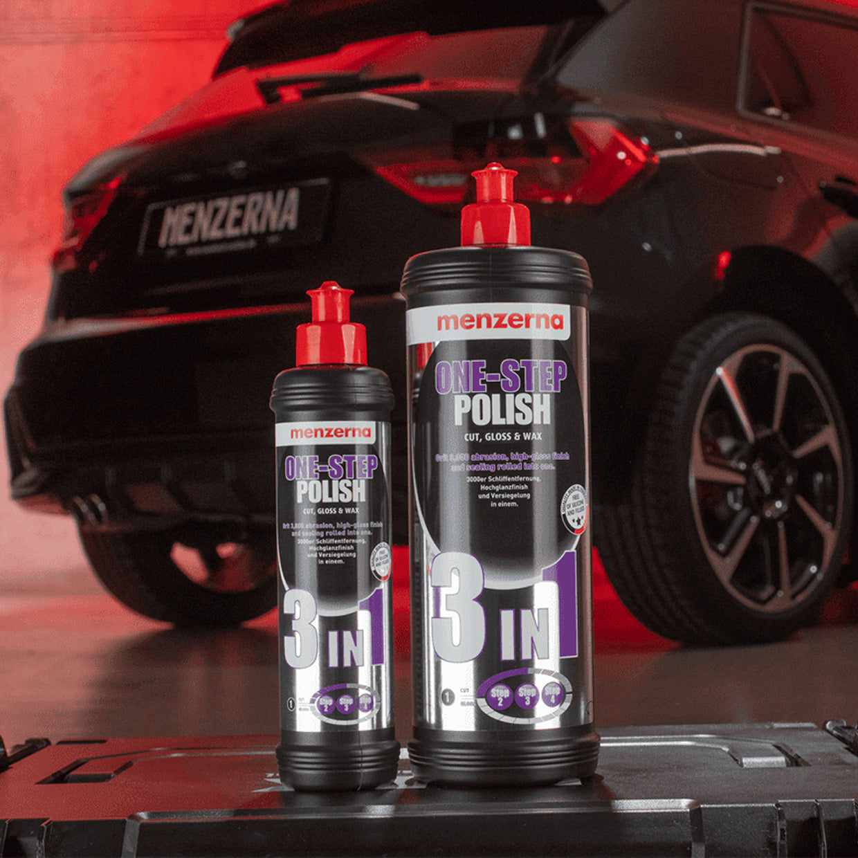 Two bottles of Menzerna One-Step Polish 3 in 1, placed in front of a car inside a garage.